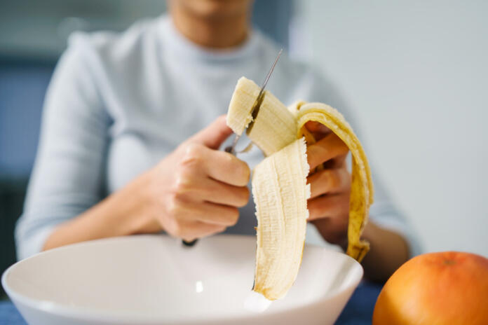 Caucasian woman sitting by the table at home cutting banana - Adult girl female preparing fruit salad at home - healthy eating concept copy space close up