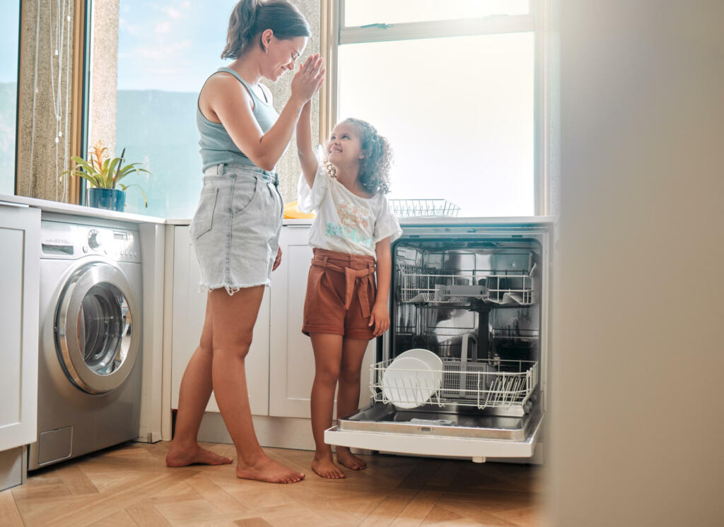 Little girl helping her mother with household chores at home. Happy mom and daughter giving high five while unloading the dishwasher together. Kid learning to be responsible by doing tasks