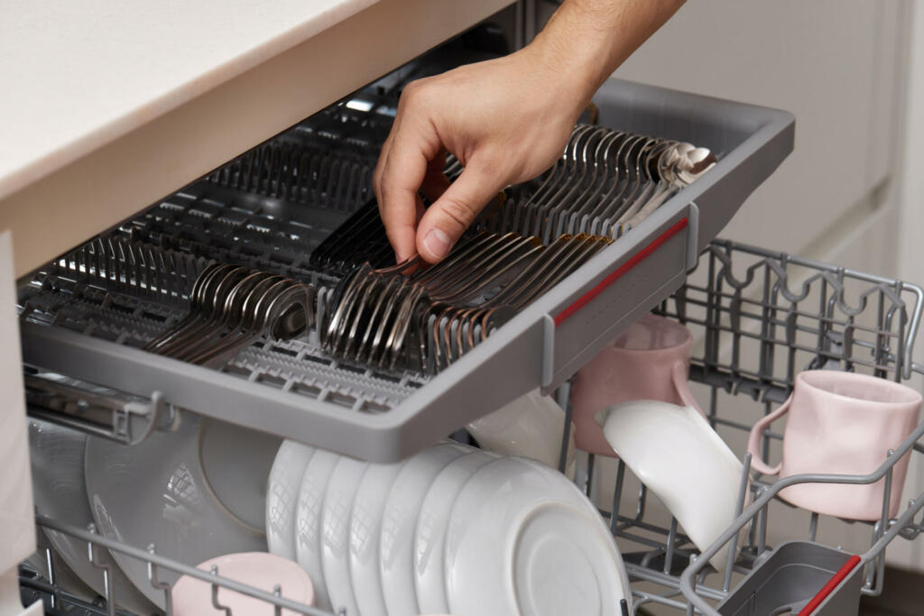 male hand unloading spoon from open automatic built-in dishwasher machine with clean utensils inside