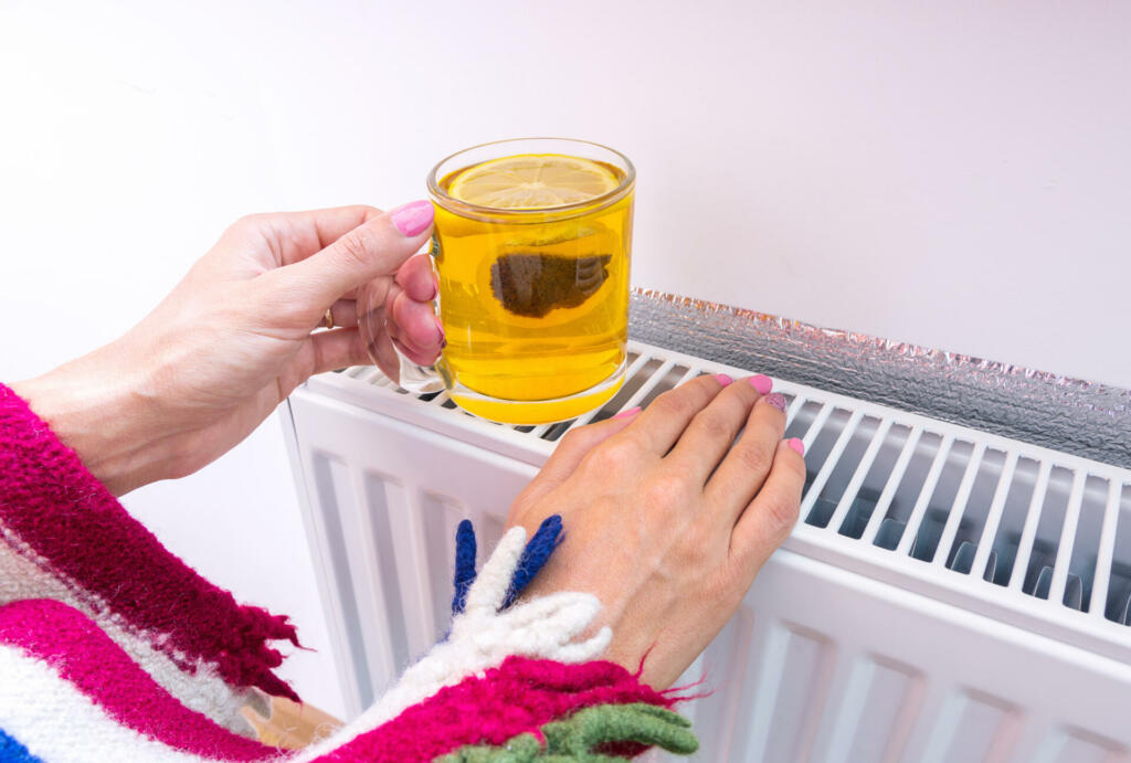 Woman warms herself near a heating radiator and drinks hot tea with lemon in the cold season. The symbolic image of the need for good heating at home in cold, autumn or winter weather.