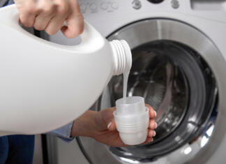 Trik za čiščenje pralnega stroja, plesen odstranimo brez težav Against background of drum of steel-colored washing machine, woman pours liquid washing gel into plastic cap. A girl in a white T-shirt carefully pours a transparent conditioner for flattening laundry