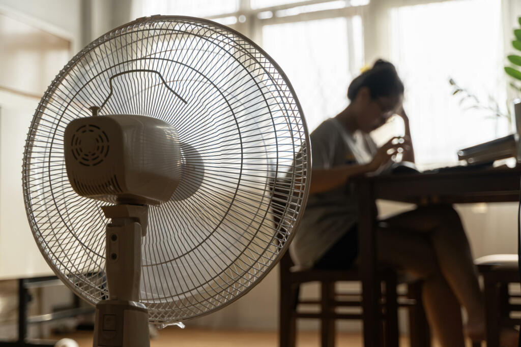 Asian unhappy woman sitting in front of working fan suffering from heat in modern house on sunny summer day in front of working fan suffering from summer heat.