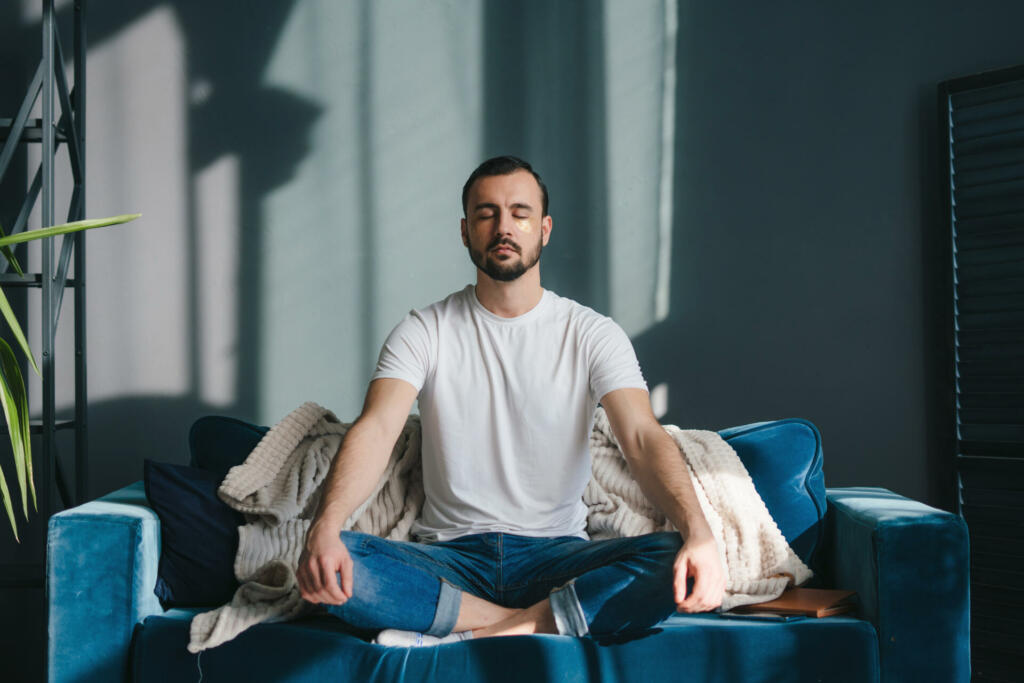 Focused man meditating in lotus pose breathing deep and slowly wearing casual clothes sitting on comfortable coach relaxing at home. Feeling free from stress.