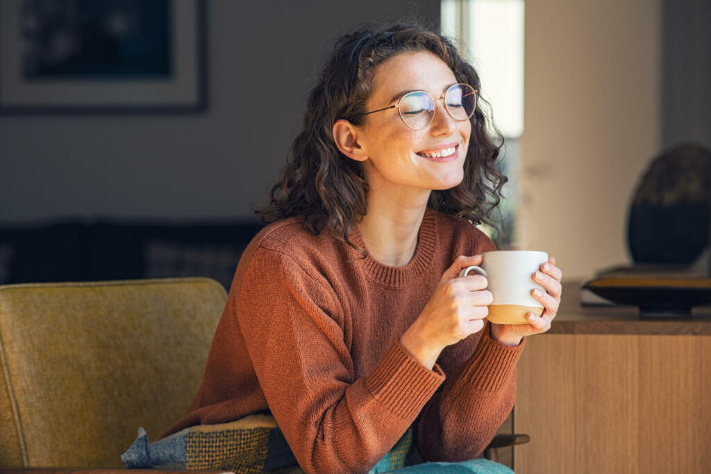 Happy young woman drinking a cup of tea in an autumn morning. Dreaming girl sitting in living room with cup of hot coffee enjoying under blanket with closed eyes. Pretty woman wearing sweater at home and enjoy a ray of sunshine on a winter afternoon.