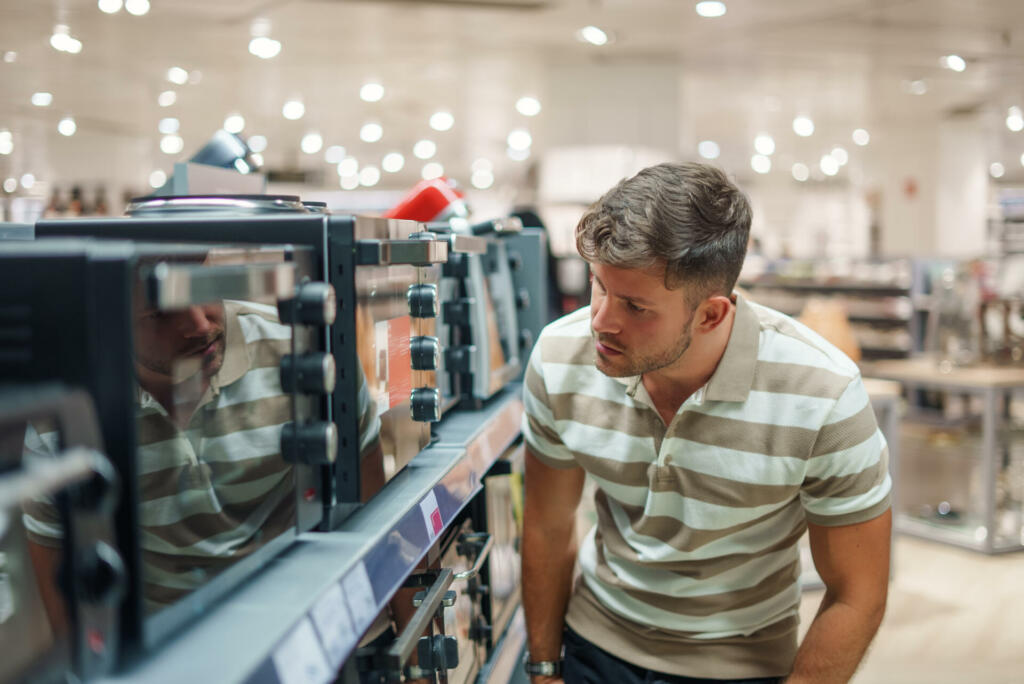 Man in casual clothes looking closely at new microwaves ovens on shelf while visiting modern mall