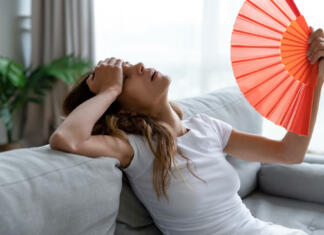 Kaj se zgodi, če imate doma pretoplo? Side view exhausted young biracial girl using paper waver, suffering from hot summer weather or high temperature at home. Unhappy overheated millennial mixed race woman feeling uncomfortable indoors.