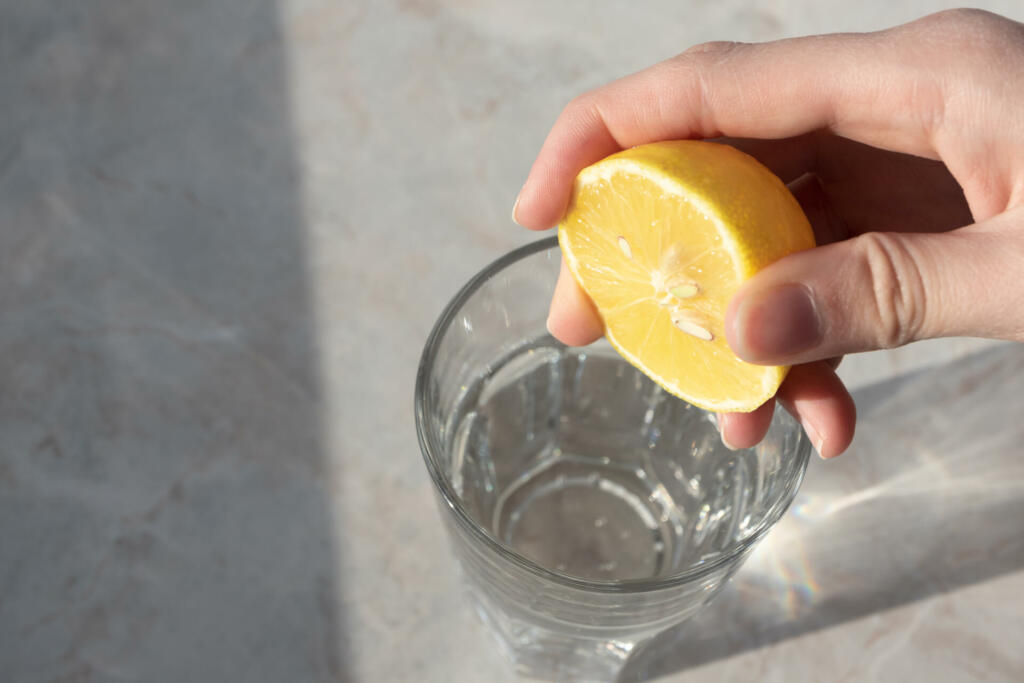 Women hand squeezes lemon juice in glass with soda water on marble table background. Detox healthy drink concept