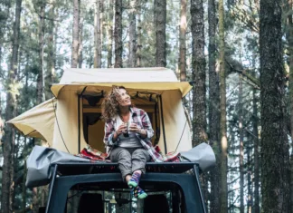 Preurejanje avtomobilov v mikro kamperje: Kateri modeli so najbolj primerni? Happy cheerful beautiful people caucasian adult woman sit down on the roof of the car with tent in background and forest around - concept of travel and enjoy nature