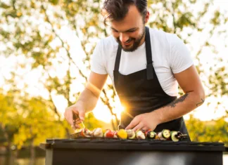 Vodič: kakšen grill žar naj kupim v 2024: oglje, plin ali električni? Crop cheerful young ethnic bearded male chef in apron preparing delicious veggie kebabs on grill and smiling during picnic in nature on sunny day