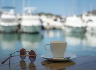 Sezona se bliža, Hrvati ‘nabijajo’ cene in pravijo še huje bo! Kepica sladoleda 3,5 €, ražnjiči 19 €, pravi šok je kava White cup with saucer, sunglasses on a table on the background of blurred boat harbor. Summer vacation concept.