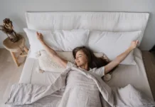 Utrujeni po 8 urah? Problem včasih ni v vas, ampak v prostoru Young long haired Italian girl laying on bed eyes closed awakening at Sunday morning smiles satisfied by healthy sleep. Top view on young woman spreading hands, stretching. Cozy home, happy people.