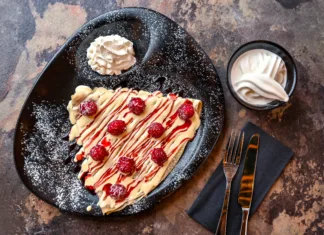 Palačinke skozi čas: izvor, zgodovina in priljubljenost po svetu in v Sloveniji White Chocolate And Raspberry Crepe with whipped cream, knife and fork served in dish isolated on dark background closeup top view of cafe baked dessert food