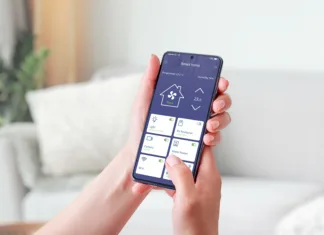 Kako vam pametni dom lahko prihrani denar? Woman's hand holding a mobile phone, showcasing a smart home app. Smart living concept. Decorated living room in background