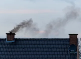 Pozor, nove obveznosti za lastnike peči! A roof with two chimneys on it. In one of the chimneys there is a pipe mounted on it were a lot of black polluting smoke is coming out or rising up from, connected to the fireplace of the house.