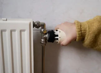 Znižajte stroške ogrevanja: trije triki za učinkovitejše delovanje radiatorjev Hand turning off the heating, radiator while wearing a warm yellow sweater. Concept of saving electricity and heating as a way of energy saving consumption.