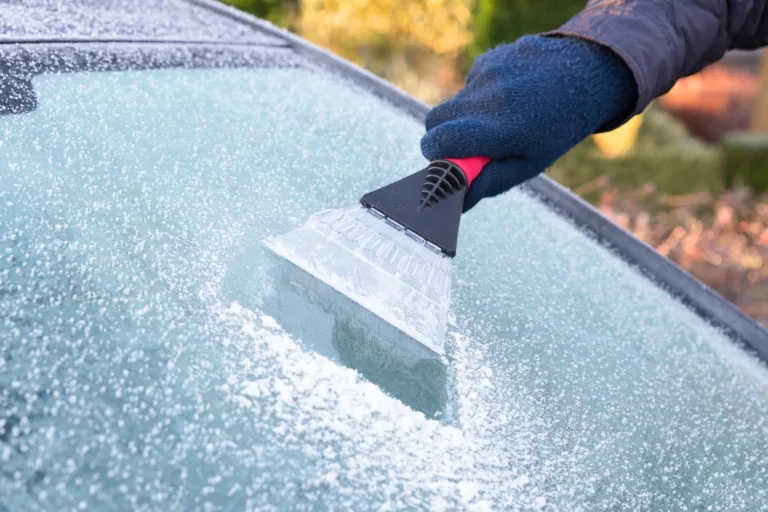 Hand wearing blue glove scratching ice from car window. The ice scraper is an extension of the arm. Part of the windshield is ice free. Only part of the window is visible. Symbol or concept of winter season, cold, frost, weather, sight.