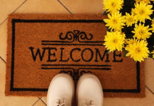 Oblika in barva predpražnika določata usodo stanovalcev! S feng šuj trikom privabite bogastvo Door mat with word Welcome, stylish boots and beautiful flowers on floor, flat lay