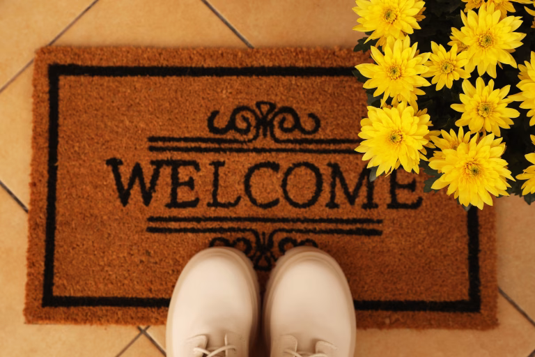 Door mat with word Welcome, stylish boots and beautiful flowers on floor, flat lay