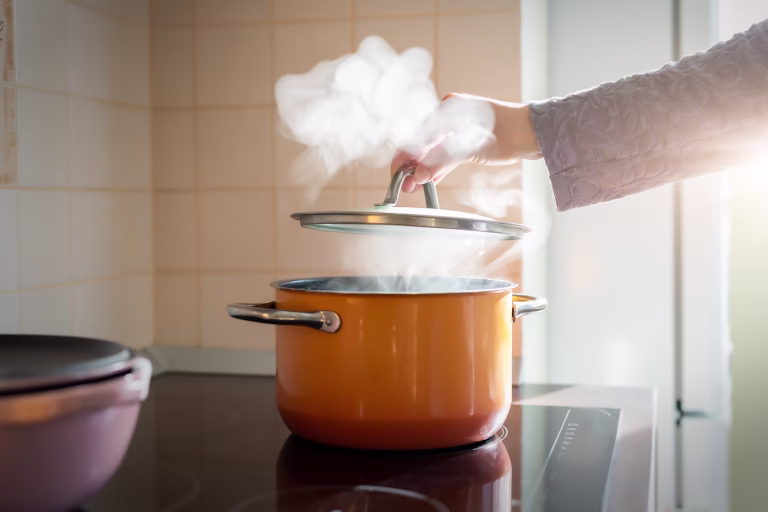 Female hand open lid of enamel steel cooking pan on electric hob with boiling water or soup and scenic vapor steam backlit by warm sunlight at kitchen. Kitchenware utensil and tool at home background.