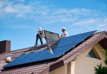 Zakaj se odločiti za samooskrbo s sončno energijo in na kaj paziti? Workers building solar panel system on roof of house. Two men installers in helmets carrying photovoltaic solar module outdoors. Alternative, green and renewable energy generation concept.