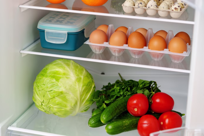 A white refrigerator is open, revealing a well-stocked interior with fresh fruits, vegetables, eggs, and other groceries.