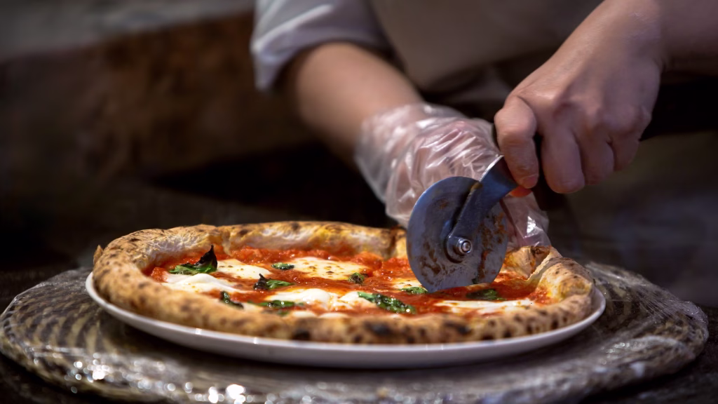 A chef is slicing a freshly baked pizza on a metal tray. The delicious meal is being prepared with precision in the professional kitchen, traditional Italian cuisine.