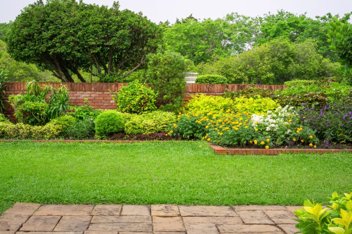 Backyard English cottage garden, colorful flowering plant and green grass lawn, brown pavement and orange brick wall, evergreen trees on background, in good care maintenance landscaping in park