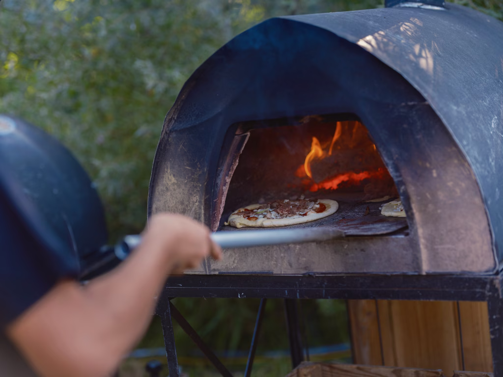 Close-up of a portable outdoor pizza oven with glowing hot flames visible inside as two pizzas cook on the stone floor. A person's arm is visible in the foreground, using a pizza peel to position or check the cooking pizzas. The charcoal-colored dome oven creates a rustic outdoor cooking scene.
