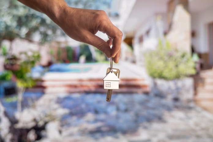 Hand holding house keys with house shaped keychain in front of a blurred background representing a new home with a swimming pool, symbolizing real estate investment and homeownership