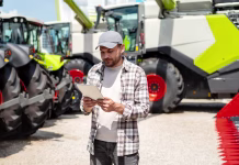 Objavljen 30-milijonski razpis za naložbe v kmetijske proizvode Man using digital tablet at agricultural machinery dealership. Farmer stands in front of new farm tractors and combine harvester.