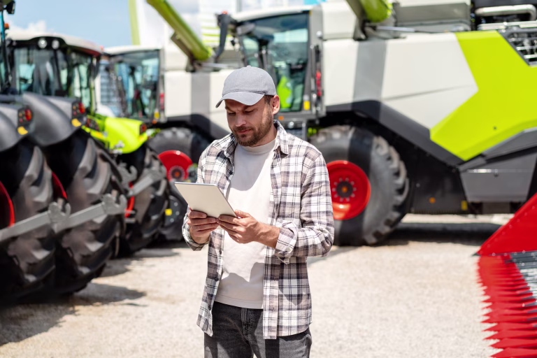 Man using digital tablet at agricultural machinery dealership. Farmer stands in front of new farm tractors and combine harvester.