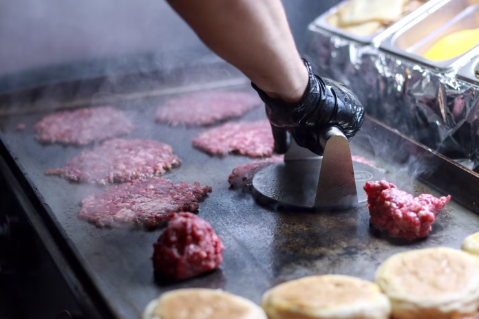 Unrecognizable person smashing raw smash burgers balls on a hot grill to make burgers in a restaurant kitchen