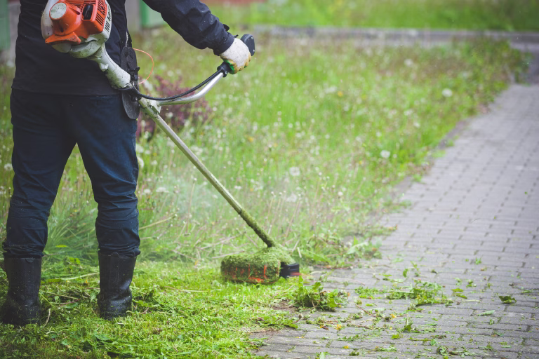 Worker in dark protective clothing with a gas mower in his hands, mowing grass in front of the house. A man mows grass with dandelions on a rainy spring day. Trimmer in the hands of a man.