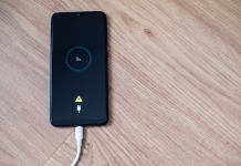 Strokovnjak opozarja: Telefona nikakor ne smete polniti ponoči, delate veliko napako! White charging cable plugged into smartphone displaying 0% battery on wooden table, emphasizing urgent need for power in modern life. Essential connection for daily communication