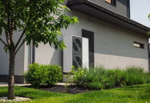 Toplotne črpalke in njihovi aduti: sodobna rešitev za ogrevanje prihodnosti Modern heat pump unit installed next to a residential house wall, surrounded by green lawn, bushes, and young trees in a landscaped yard. High quality photo