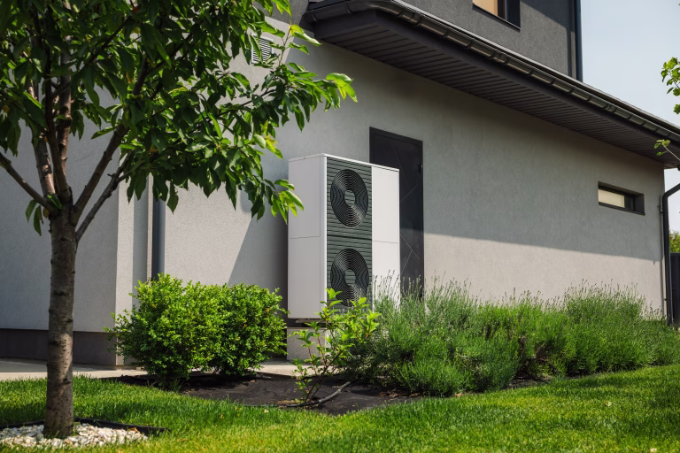 Modern heat pump unit installed next to a residential house wall, surrounded by green lawn, bushes, and young trees in a landscaped yard. High quality photo