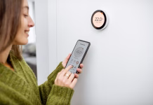 Energijska pamet doma: povezani sistemi za maksimalni izkoristek toplote Woman dressed in green sweater regulating heating temperature with a modern wireless thermostat and smart phone at home. Synchronization of thermostat with mobile devices concept