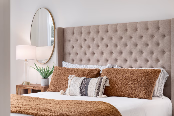 A bedroom detail shot with a cushioned headboard, a gold mirror on the wall, and a plant and lamp on the nightstand.