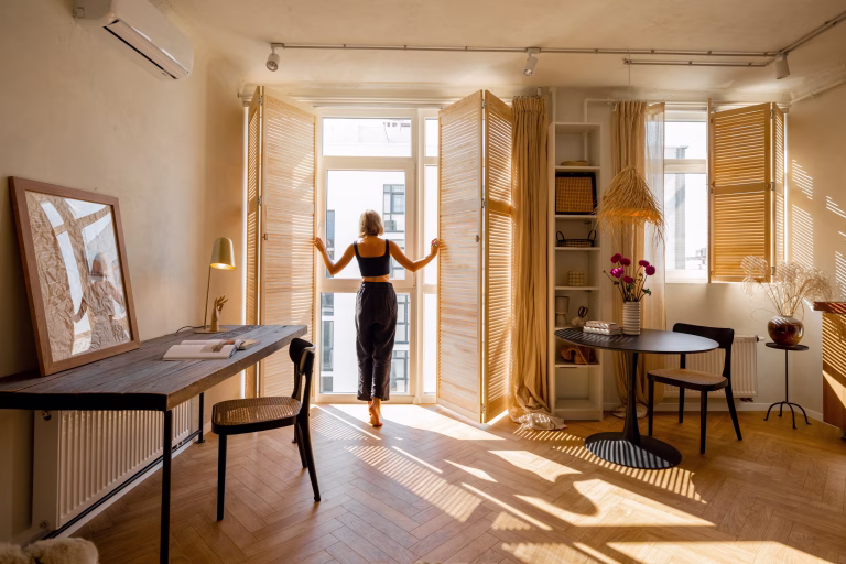 Woman opens window blinds letting the sun inside the room, spending good morning in sunny and cozy apartment in beige tones. Interior view