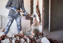Ali je zid nosilen? Kako oceniti nosilnost zidu, da ne bo napak Close up of sledgehammer blow on brick, plaster. Workman striking devastating blow at remnants of interoom wall against backdrop of table and other plastered walls.