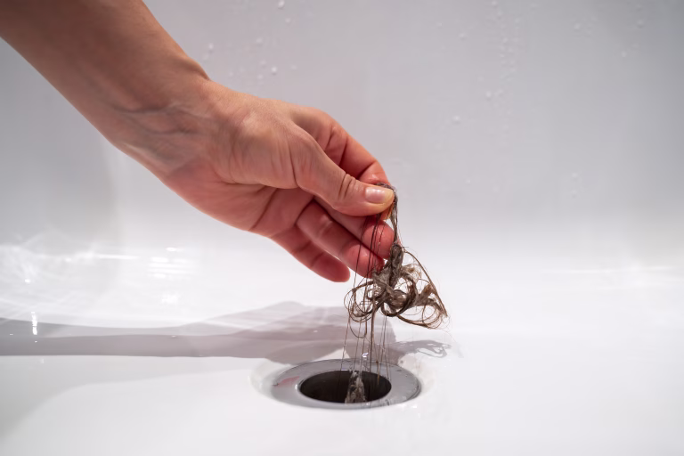 Hand pulls hair out of bathroom drain. Female palm extracts shock of hair from siphon in bathroom. Water drain hole clogged with hair clump. Close up. Cleaning shower polluted sewerage system.