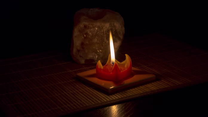 The flame of a red candle illuminates the stone salt tea light lamp on on a bamboo mat with black background.