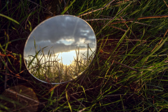 A round mirror lies on a grassy field, reflecting a cloudy sky with the sundown.