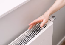 Švedska metoda za brezplačno ogrevanje: fantastična ideja, ki rešuje visoke račune A hand checking warmth of a new electric heater in a cozy apartment. Well designed and modern white radiator ensures comfortable warmth during cold winter days while enhancing the room aesthetics.
