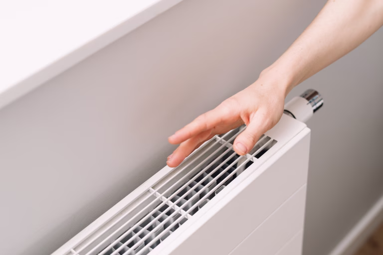 A hand checking warmth of a new electric heater in a cozy apartment. Well designed and modern white radiator ensures comfortable warmth during cold winter days while enhancing the room aesthetics.