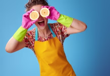 Čisto gospodinjstvo s pomočjo … limone Big cleaning time. smiling modern housewife in a yellow apron holding half a lemon in front of eyes isolated on blue