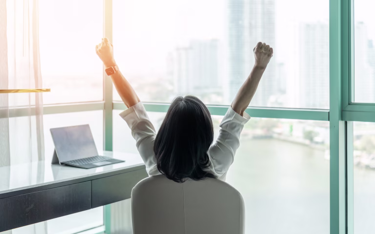 Business achievement concept with happy businesswoman relaxing in office or hotel room, resting and raising fists with ambition looking forward to city building urban scene through glass window