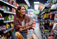 Zakaj iz trgovine skoraj nikoli ne pridemo samo s kruhom? Poskusite 30-dnevni izziv, ki lahko spremeni vaše finance Happy woman using grocery list while shopping at supermarket.