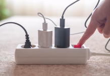 Zakaj se vam aparati pokvarijo že po dveh letih? To napako delajo skoraj vsi, a je lahko zelo draga … Person pressing red on/off switch on white power strip to cut electricity to multiple chargers, symbolizing mindful power use, energy saving, lower bills and household sustainability