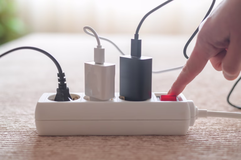 Person pressing red on/off switch on white power strip to cut electricity to multiple chargers, symbolizing mindful power use, energy saving, lower bills and household sustainability